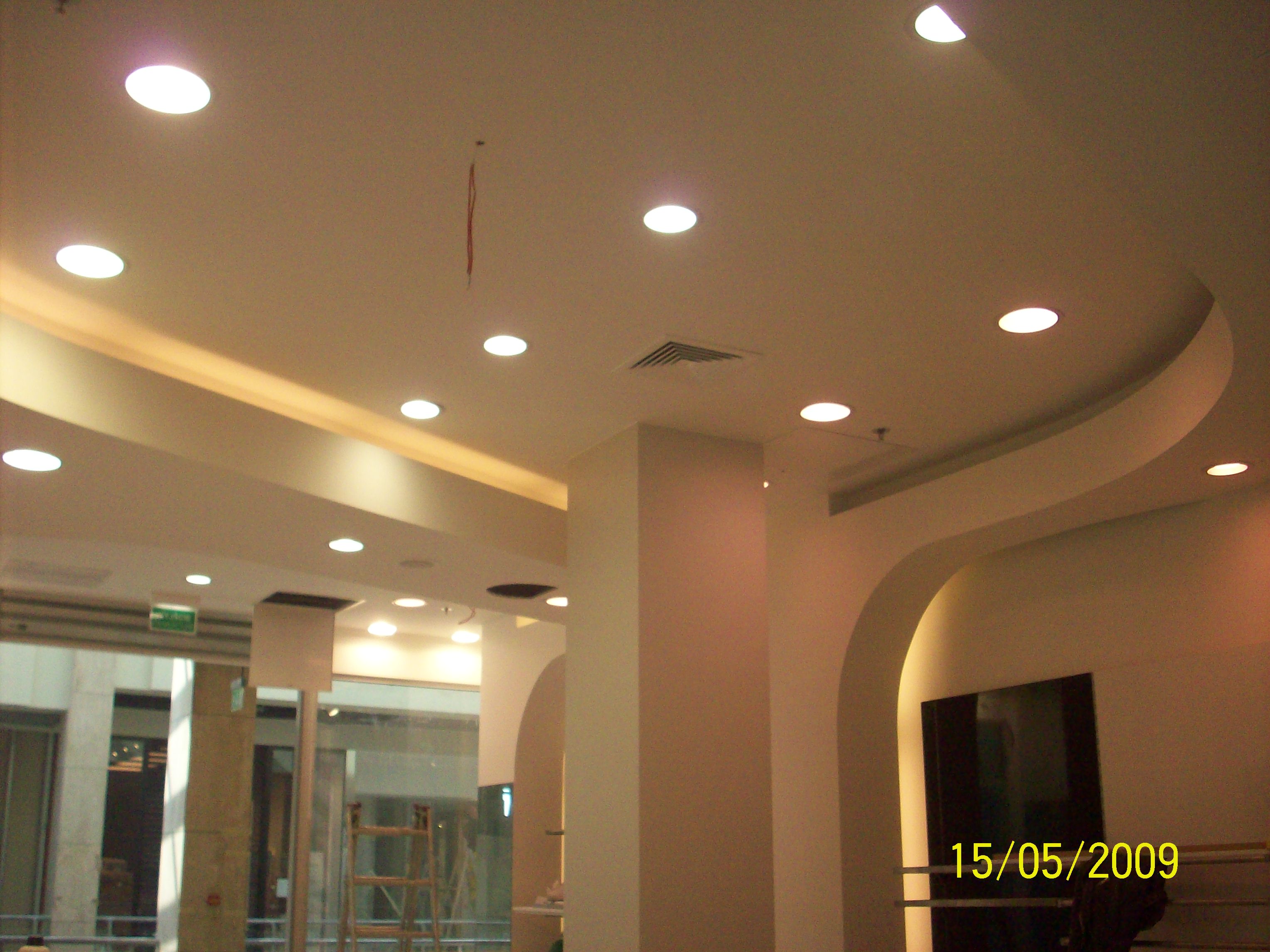 Sufit z zamontowanymi okrągłymi lampami halogenowymi, widoczny fragment instalacji wentylacyjnej, kolumna i łuk w jasnym odcieniu beżu, fragment okna z widoczną drabiną w tle.